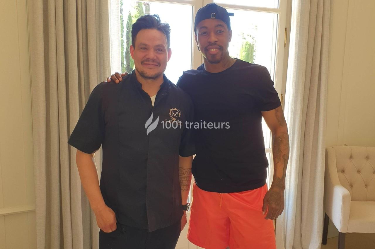 Deux hommes souriants posent côte à côte dans une pièce lumineuse devant une fenêtre avec des rideaux.