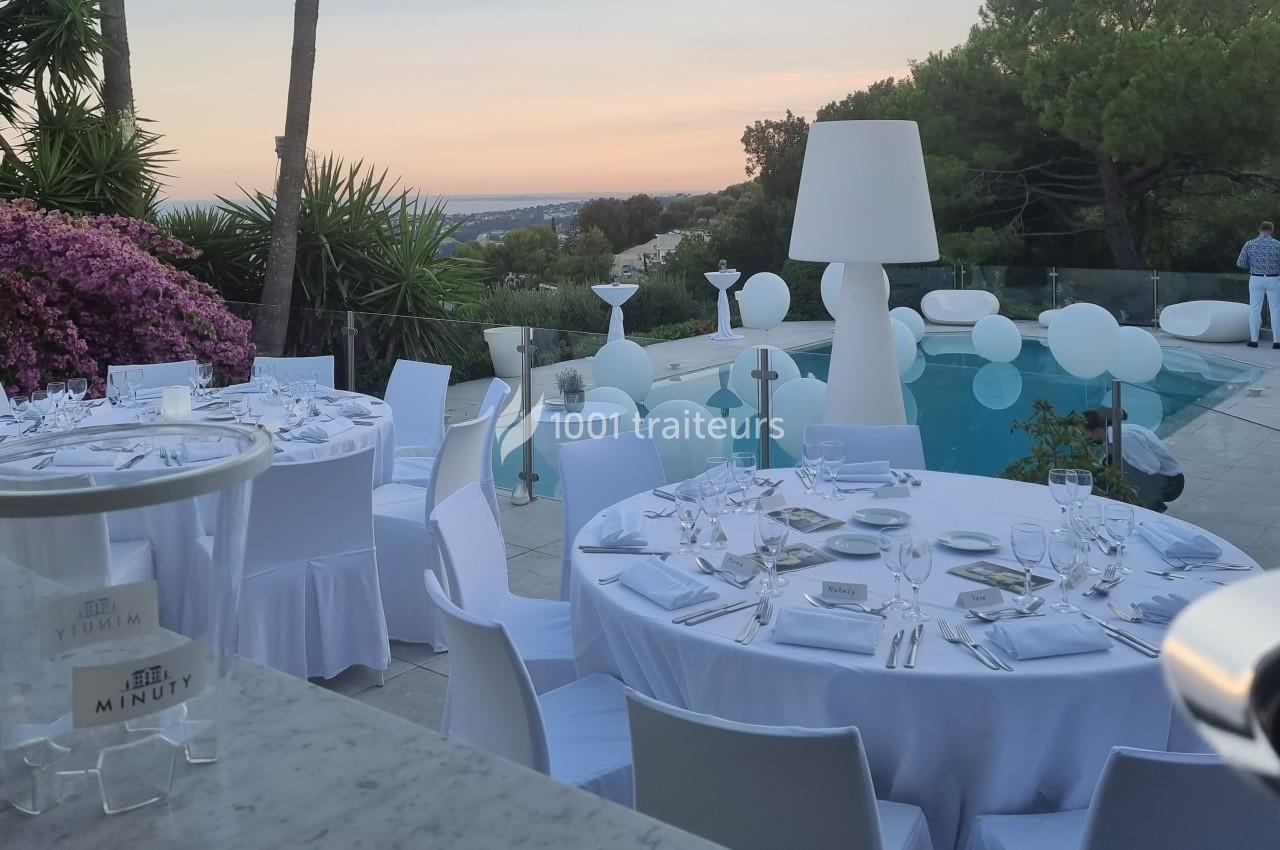 Tables dressées avec nappes blanches autour d'une piscine au coucher du soleil, décorées de ballons et de végétation.