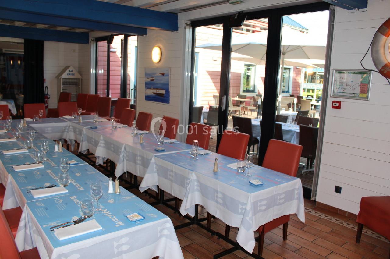 Salle de restaurant lumineuse avec tables dressées, nappes bleues et chaises rouges, donnant sur une terrasse extérieure.