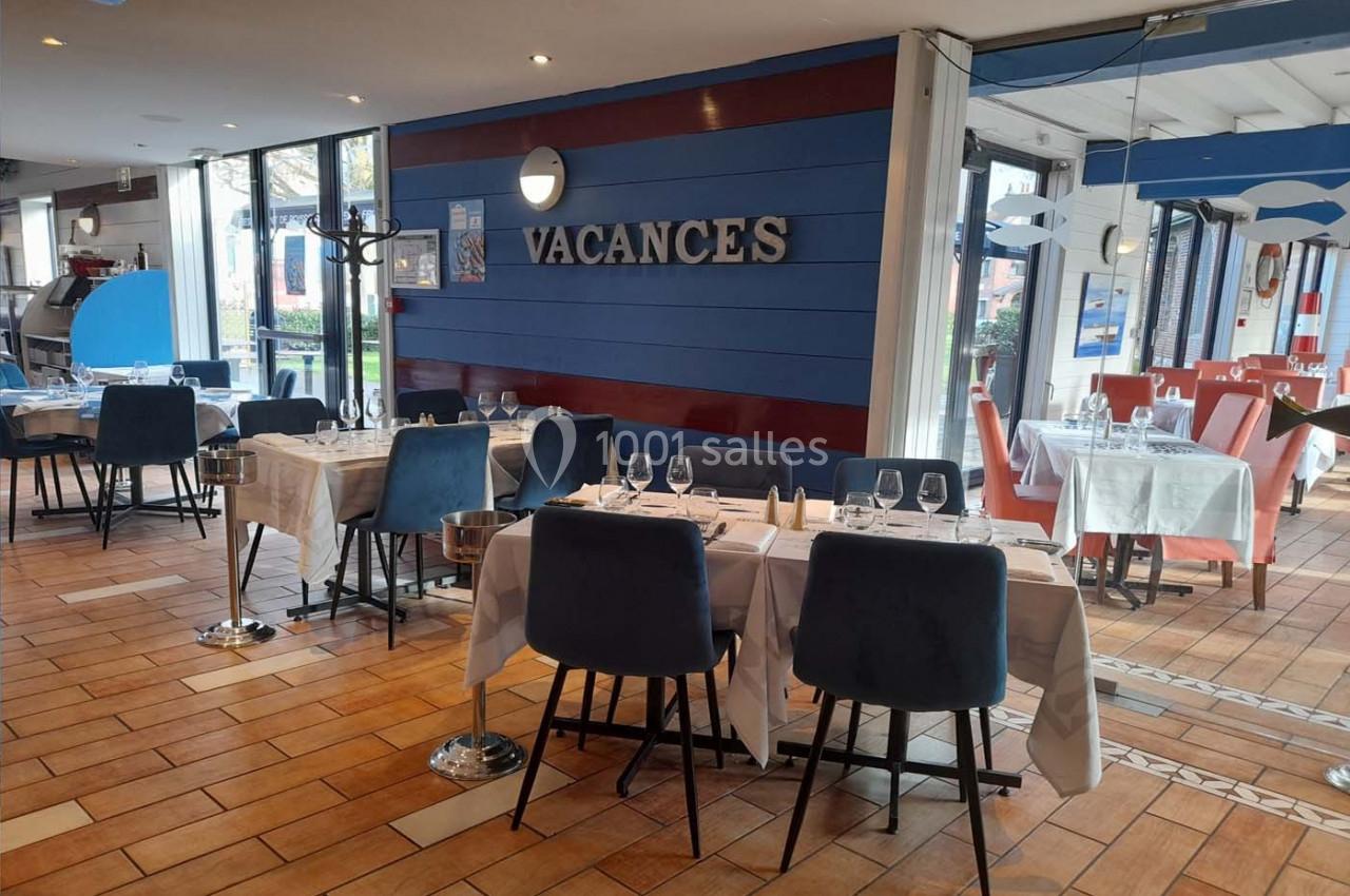 Salle de restaurant lumineuse avec tables dressées, chaises bleues et mur décoré du mot ’VACANCES’.