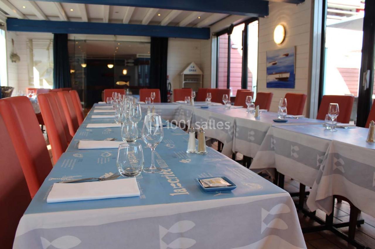 Salle de restaurant lumineuse avec de longues tables dressées, nappes bleues et chaises rouges.