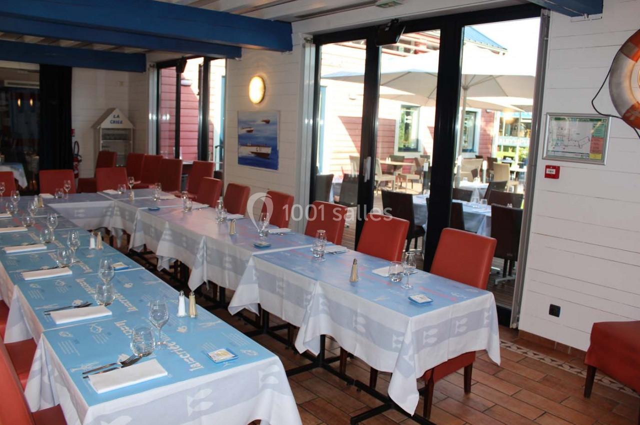 Salle de restaurant lumineuse avec tables dressées, nappes bleues et blanches, chaises rouges et terrasse visible à l…