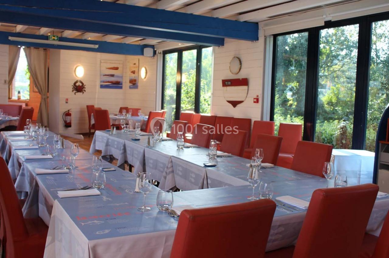 Salle de restaurant lumineuse avec tables dressées, chaises rouges et grandes baies vitrées donnant sur un jardin.