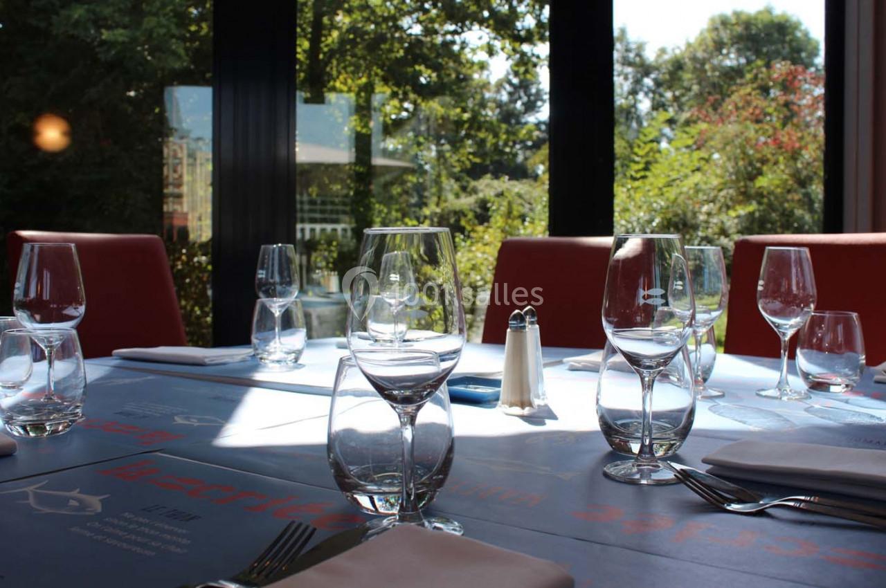 Table dressée avec des verres, couverts et serviettes, près de grandes baies vitrées donnant sur un jardin verdoyant.