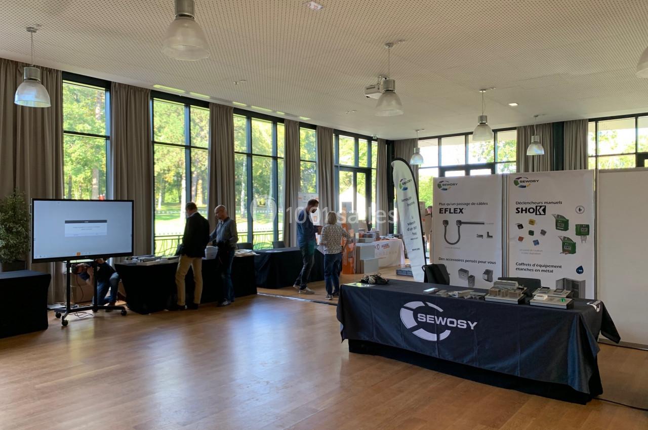 Salle lumineuse avec stands d'exposition, panneaux informatifs et visiteurs discutant près des fenêtres.