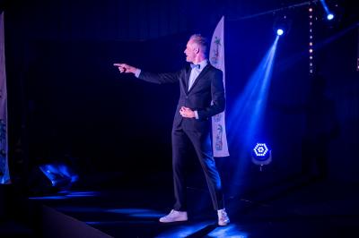 Un homme en costume noir et baskets blanches pointe du doigt sur une scène éclairée par des projecteurs.