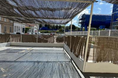 Terrasse ombragée avec des canisses, entourée de bâtiments et d'une structure bleue moderne en arrière-plan.