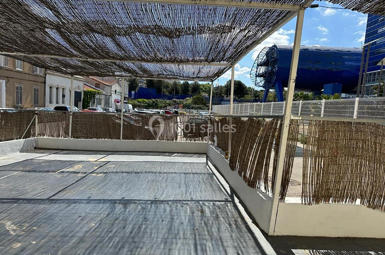 Terrasse ombragée avec des canisses, entourée de bâtiments et d'une structure bleue moderne en arrière-plan.