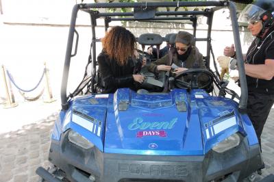Miniature Randonnée en buggy Deux personnes assises dans un buggy bleu en extérieur, discutant et manipulant des objets, avec un homme debout à côté.