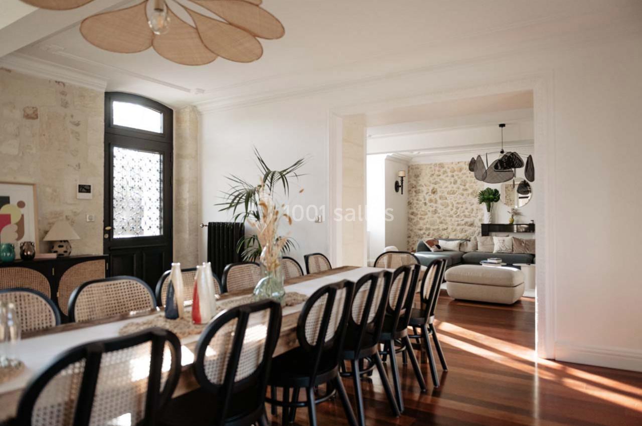 Salle à manger lumineuse avec une grande table en bois, chaises noires et vue sur un salon avec murs en pierre.