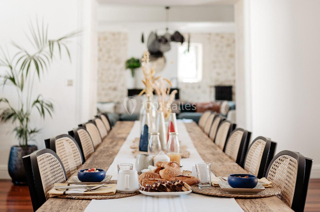 Table en bois longue avec des chaises en rotin, dressée pour un repas avec pains, fruits et carafes dans une pièce lumineuse.