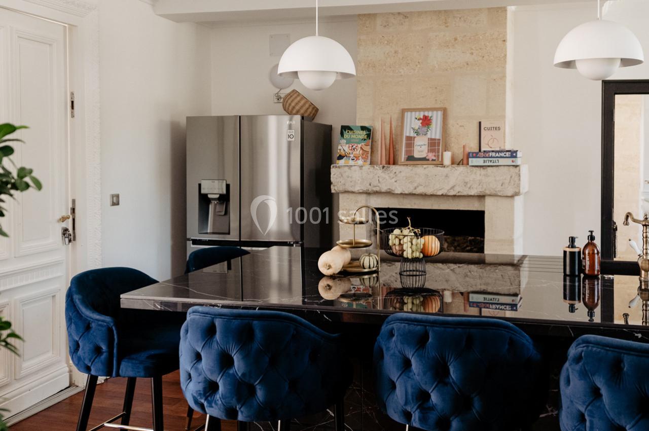 Cuisine moderne avec un îlot central en marbre noir, chaises bleues, réfrigérateur argenté et cheminée décorative.