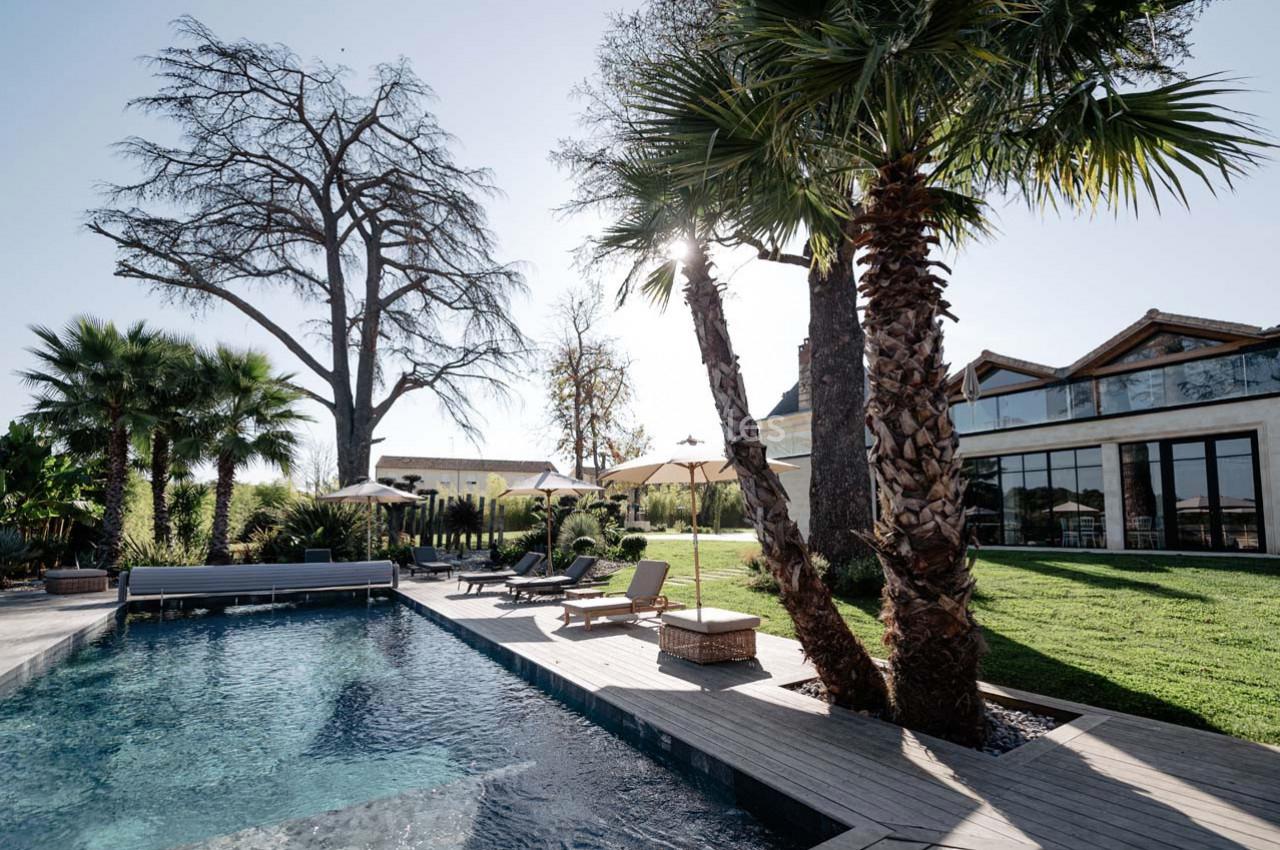 Piscine extérieure entourée de palmiers, transats et pelouse, avec un bâtiment moderne en arrière-plan.