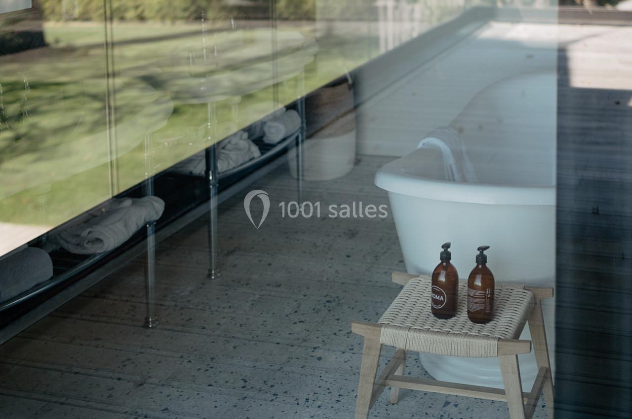 Baignoire blanche près d'une fenêtre, avec deux flacons de savon sur un tabouret en bois dans un espace lumineux.