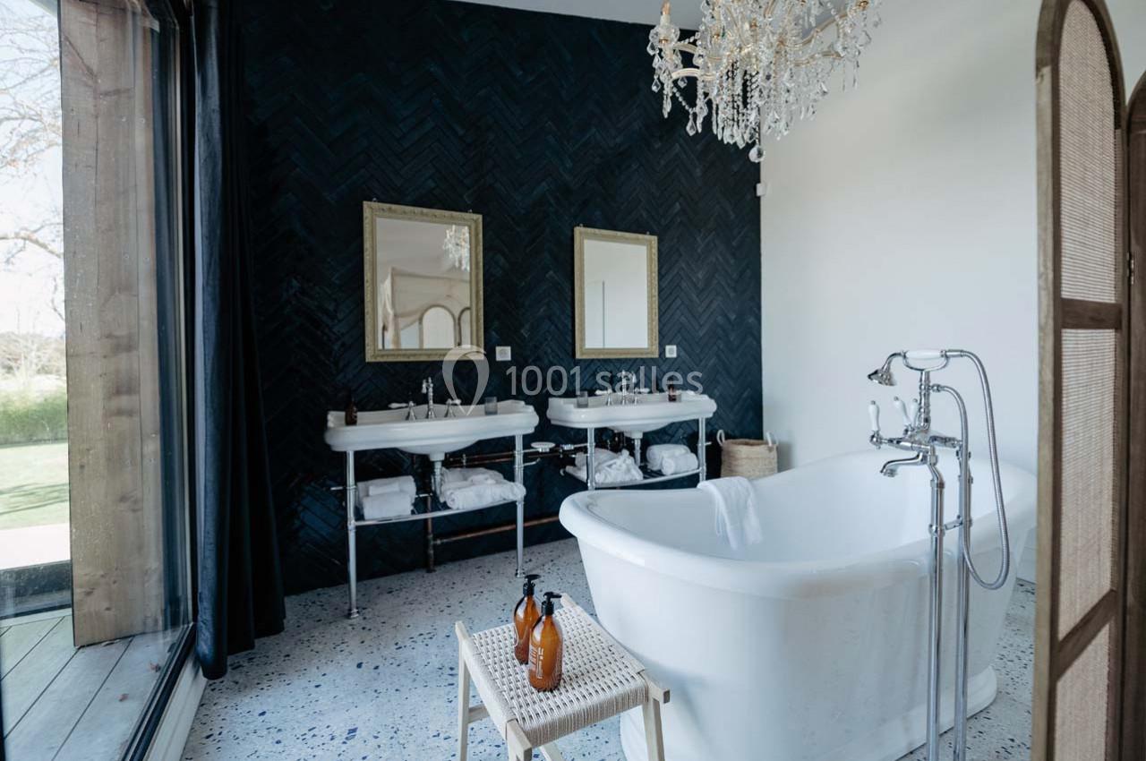 Salle de bain élégante avec baignoire blanche, double vasque, mur sombre en chevrons et lustre en cristal.
