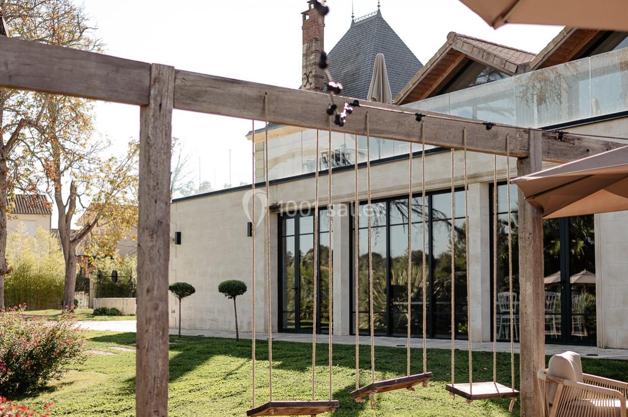 Balançoires en bois suspendues à une structure dans un jardin, avec un bâtiment moderne en arrière-plan.