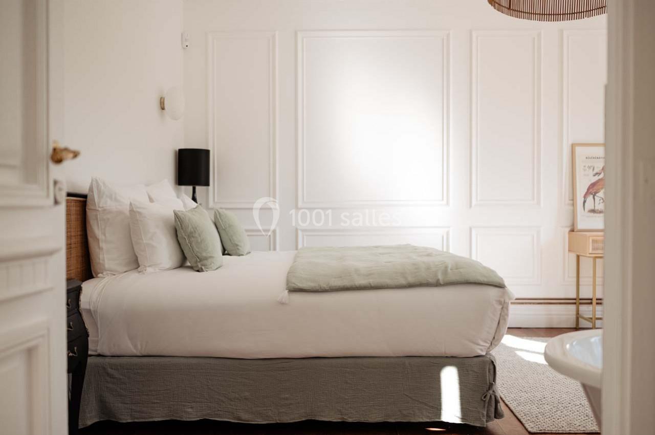 Chambre lumineuse avec lit double, draps blancs, coussins verts, murs blancs moulurés et lampe noire.