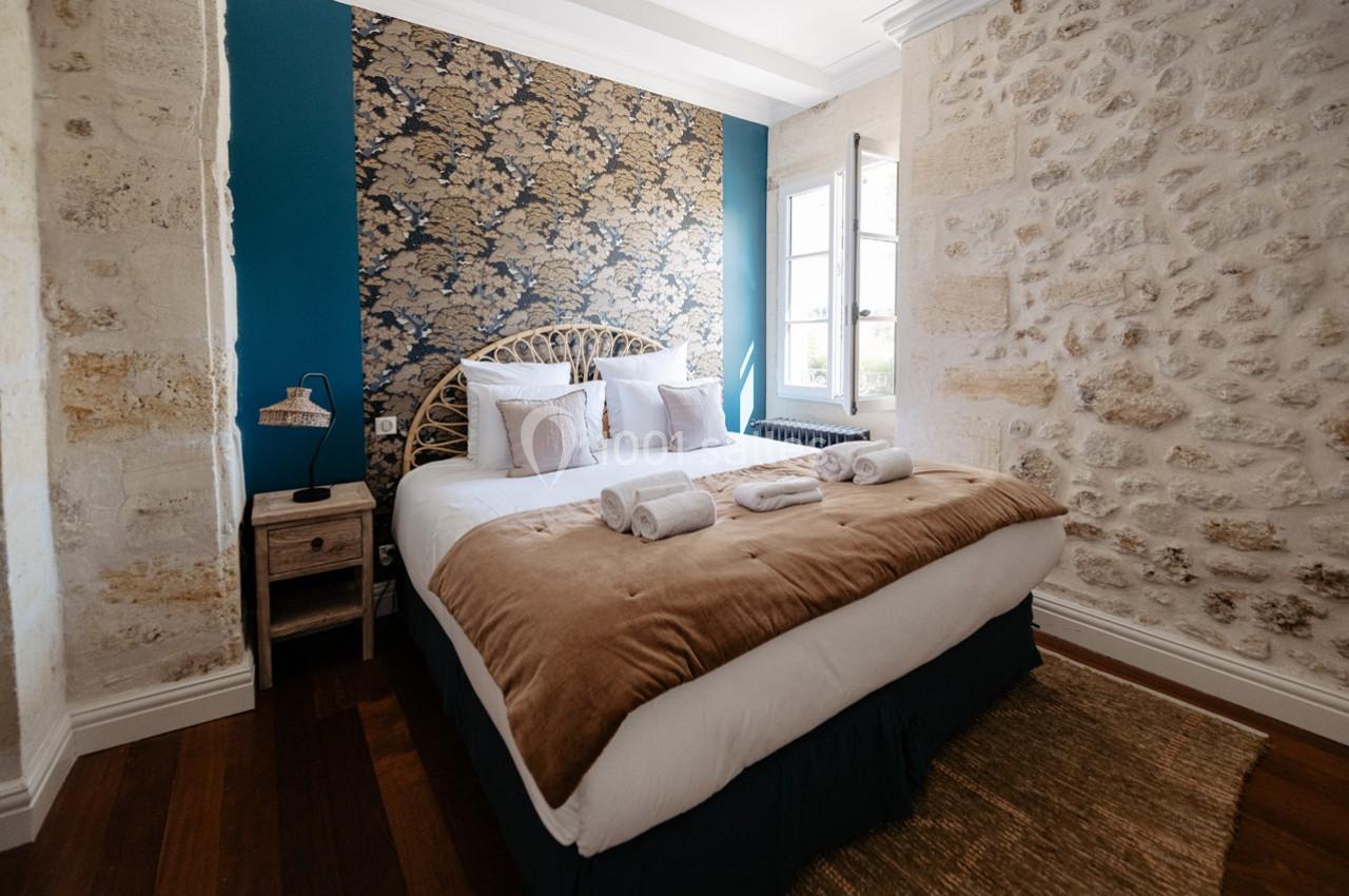 Chambre lumineuse avec lit double, murs en pierre apparente, tête de lit en rotin et fenêtre donnant sur l'extérieur.