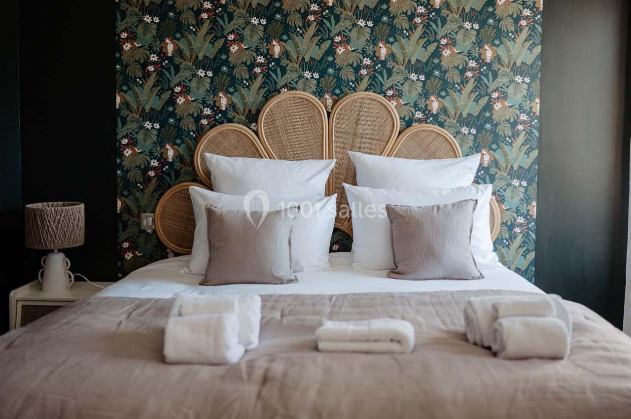 Lit double avec tête de lit en rotin, draps blancs, coussins beiges et fond de mur à motif végétal.