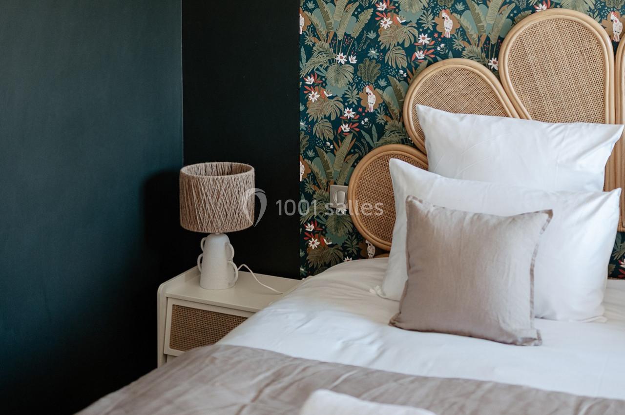 Lit avec coussins blancs et beiges, tête de lit en rotin, lampe en céramique et mur décoré de papier peint floral.