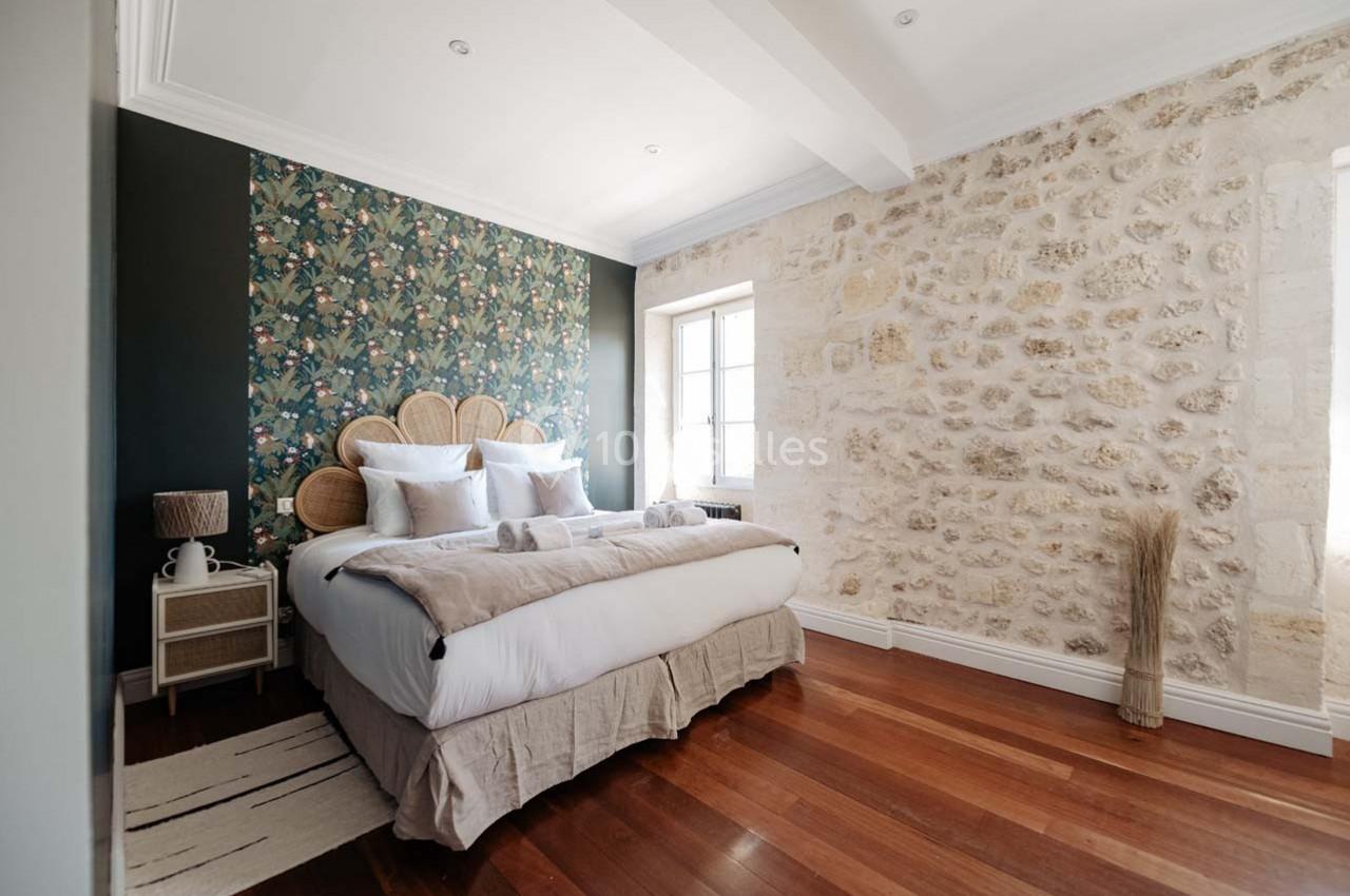 Chambre lumineuse avec lit double, tête de lit en rotin, mur en pierre apparente et parquet en bois.