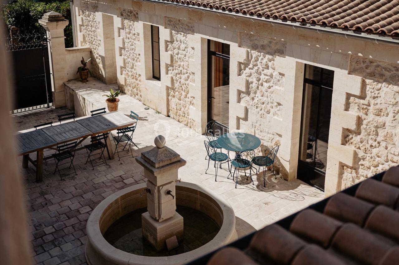 Cour en pierre avec une fontaine centrale, une grande table en bois et des chaises près d'une maison en pierre.