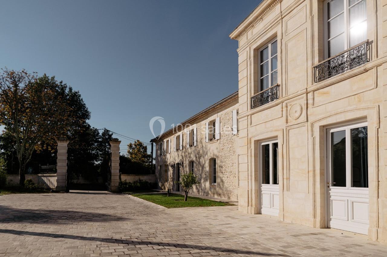 Façade d'un bâtiment en pierre avec grandes fenêtres, cour pavée et arbres sous un ciel dégagé.