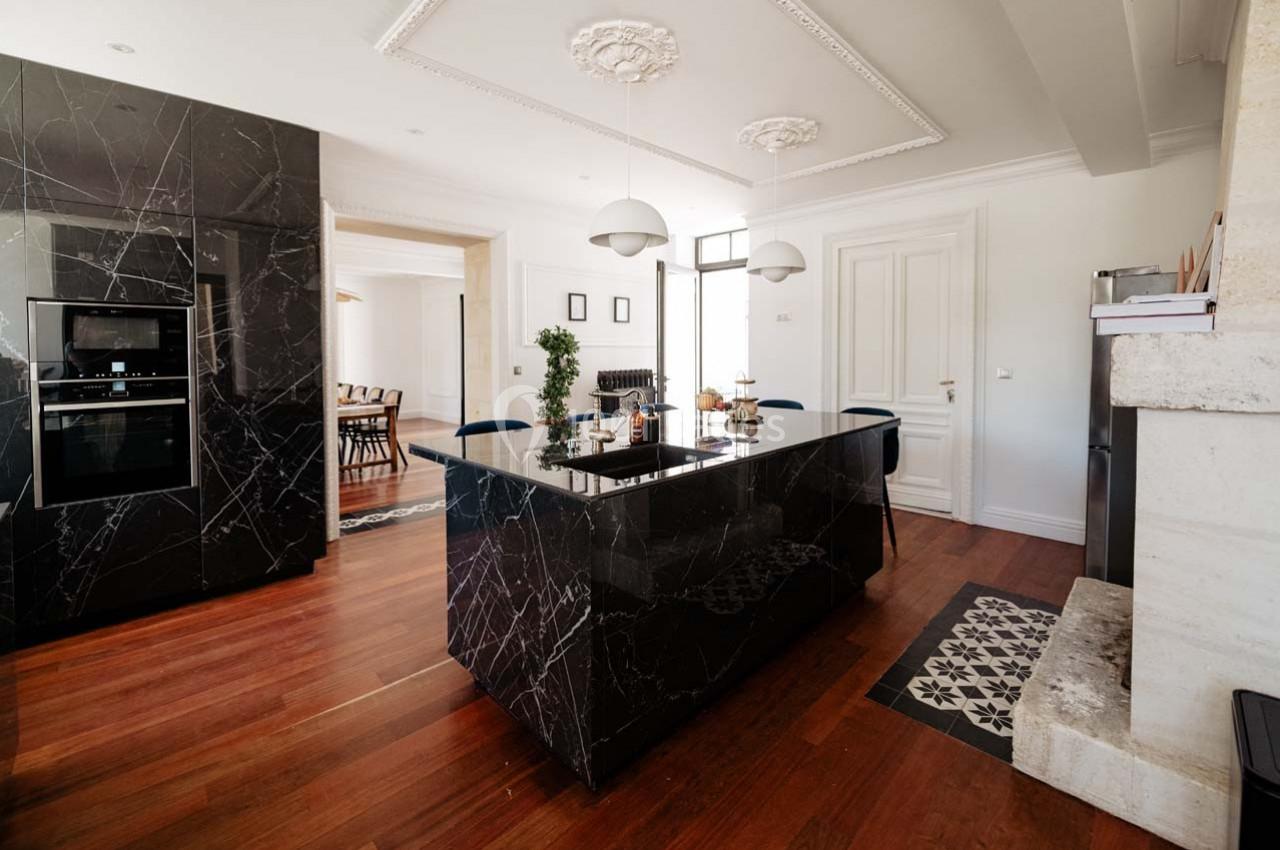 Cuisine moderne avec îlot central en marbre noir, parquet en bois, luminaires suspendus et salle à manger en arrière-plan.