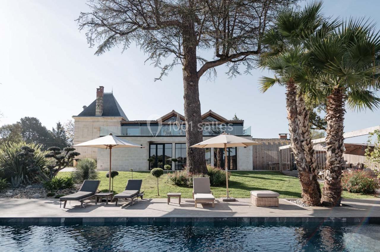 Jardin avec piscine, transats et parasols devant une grande maison en pierre entourée d'arbres.