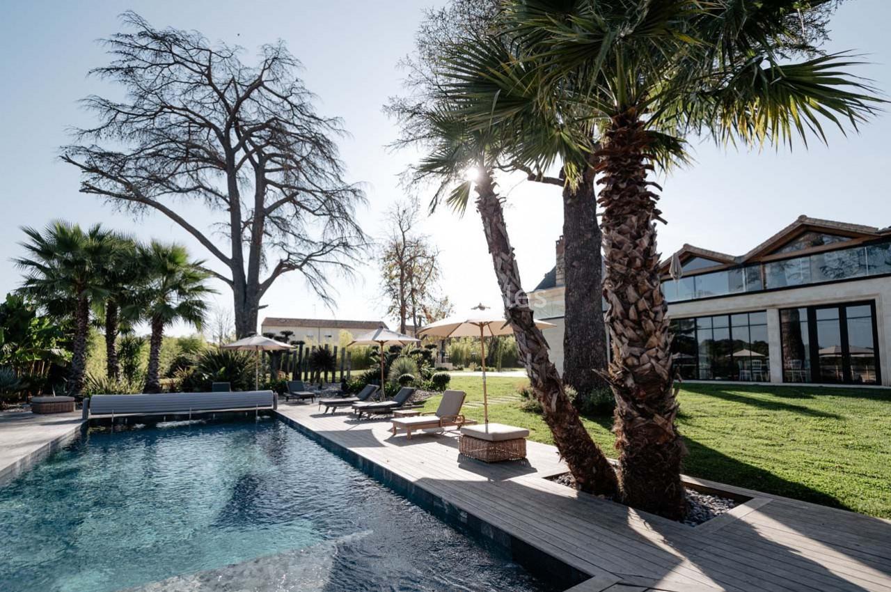 Piscine extérieure entourée de palmiers, transats et pelouse, avec bâtiment moderne en arrière-plan.