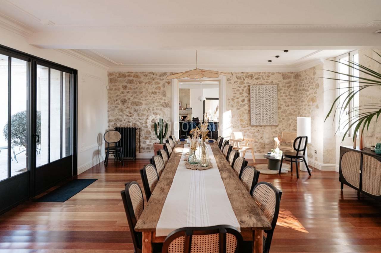 Salle à manger lumineuse avec une grande table en bois, des chaises en rotin, parquet et murs en pierre apparente.