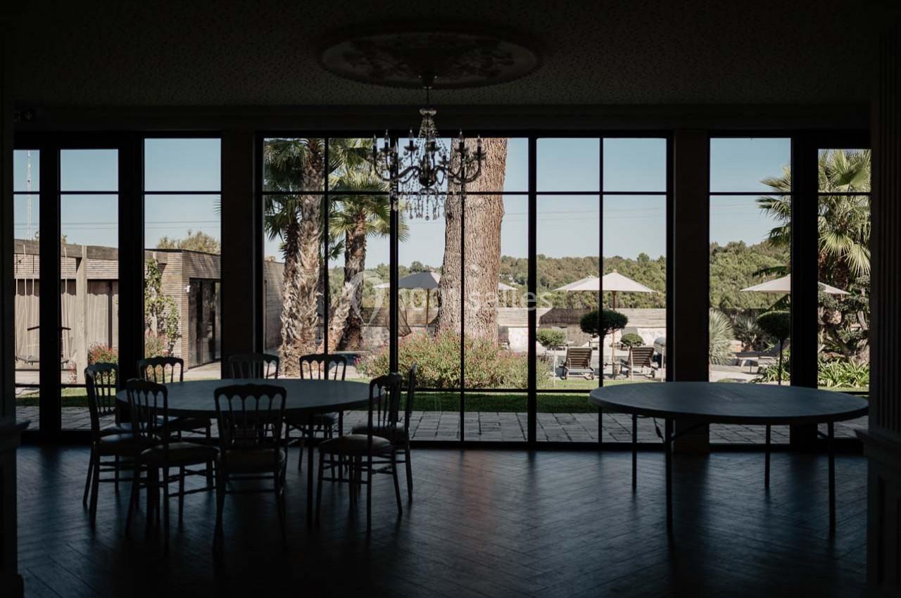 Salle sombre avec des tables et chaises devant de grandes baies vitrées donnant sur un jardin arboré et ensoleillé.