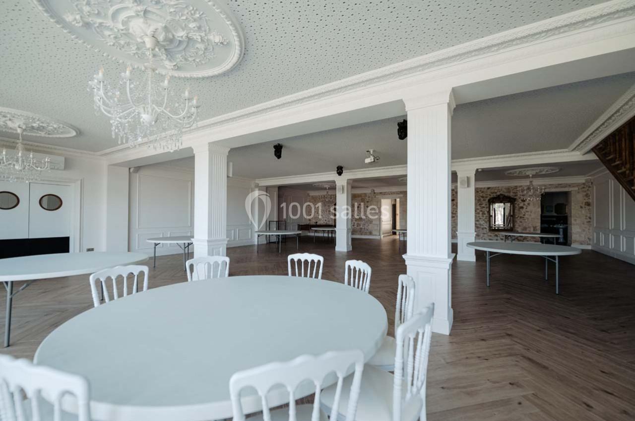 Salle lumineuse avec tables rondes blanches, chaises assorties, moulures au plafond et parquet clair.