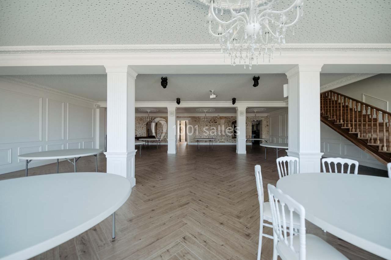Salle lumineuse avec parquet en chevrons, murs blancs, tables rondes et chaises, escalier en bois visible au fond.