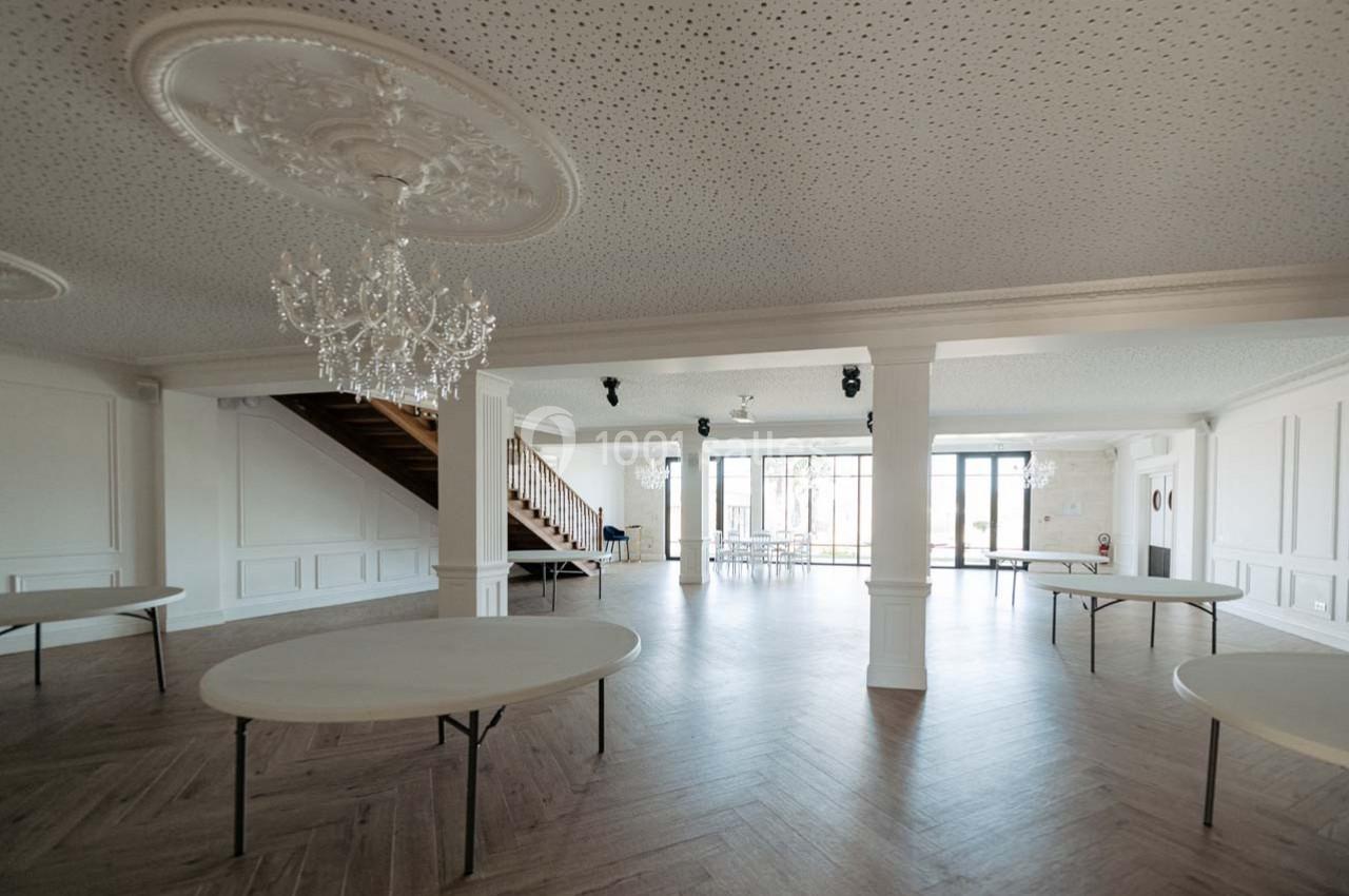 Salle lumineuse avec parquet en bois, tables rondes pliantes, lustres suspendus et escalier en bois sur le côté.