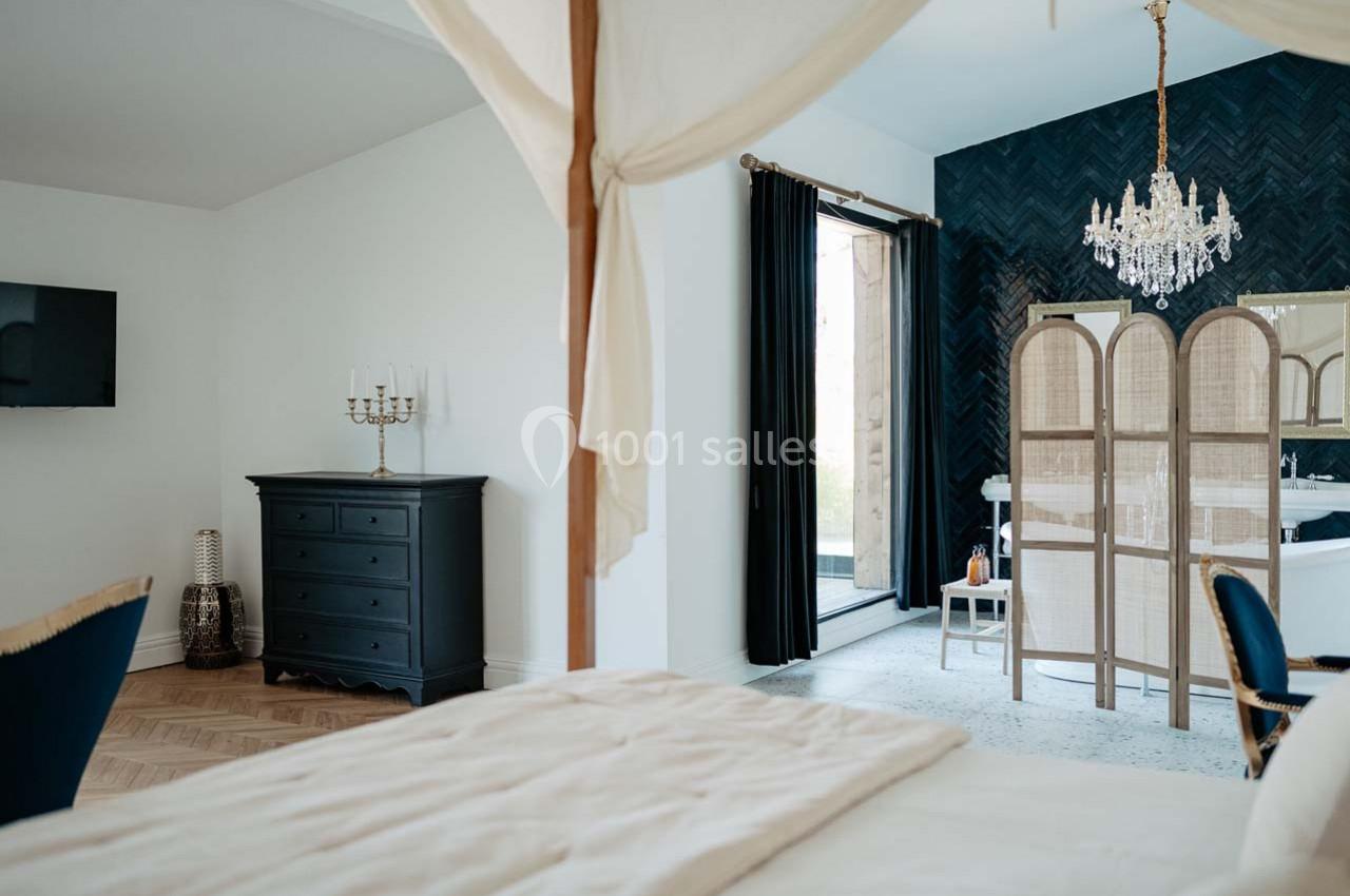 Chambre élégante avec lit à baldaquin, commode noire, paravent en rotin, lustre et accès à une terrasse.