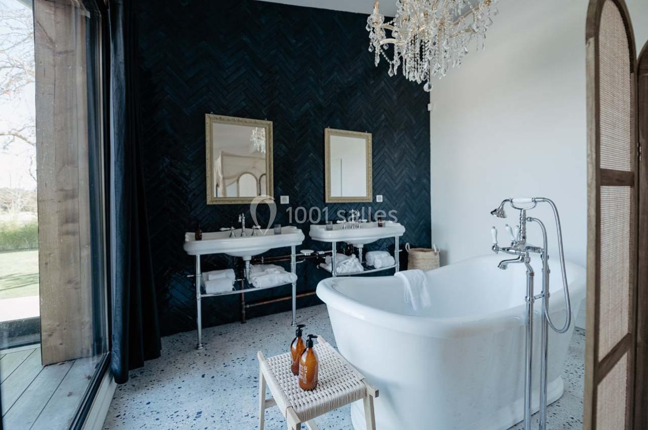 Salle de bain élégante avec baignoire blanche, double vasque, mur sombre en chevrons et lustre décoratif.