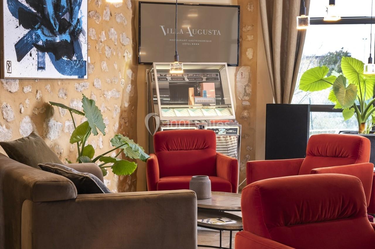 Salon avec fauteuils rouges et gris, jukebox rétro, plantes vertes et mur en pierre dans un cadre lumineux.