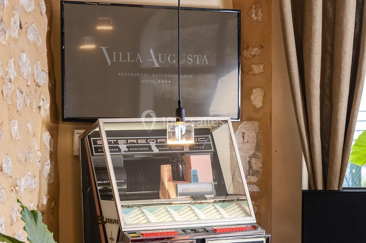 Une jukebox rétro sous une lampe suspendue, avec un écran affichant ’Villa Augusta’ en arrière-plan.