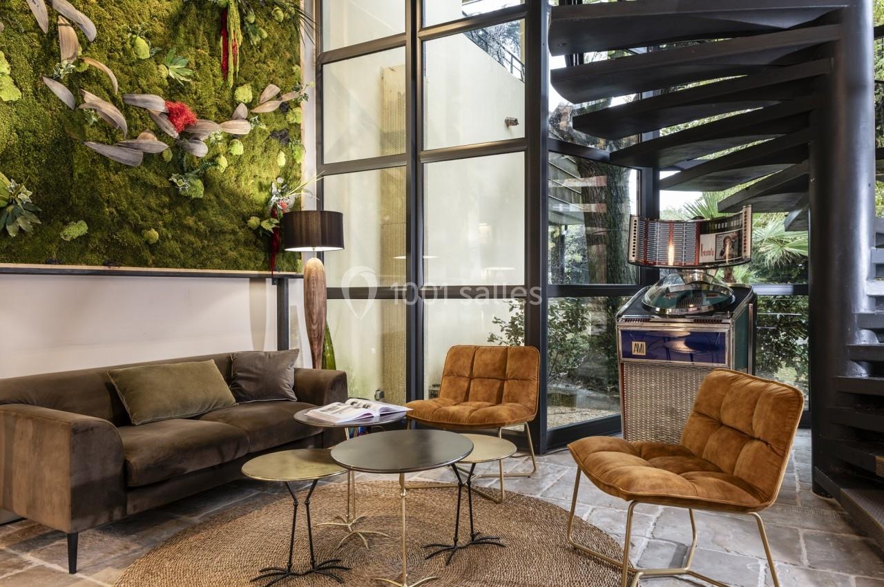 Salon moderne avec canapé, fauteuils en velours, tables basses, mur végétal, jukebox et escalier en colimaçon.