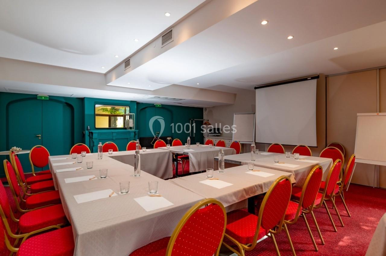 Salle de réunion lumineuse avec tables disposées en U, chaises rouges, tableau blanc et écran de projection.