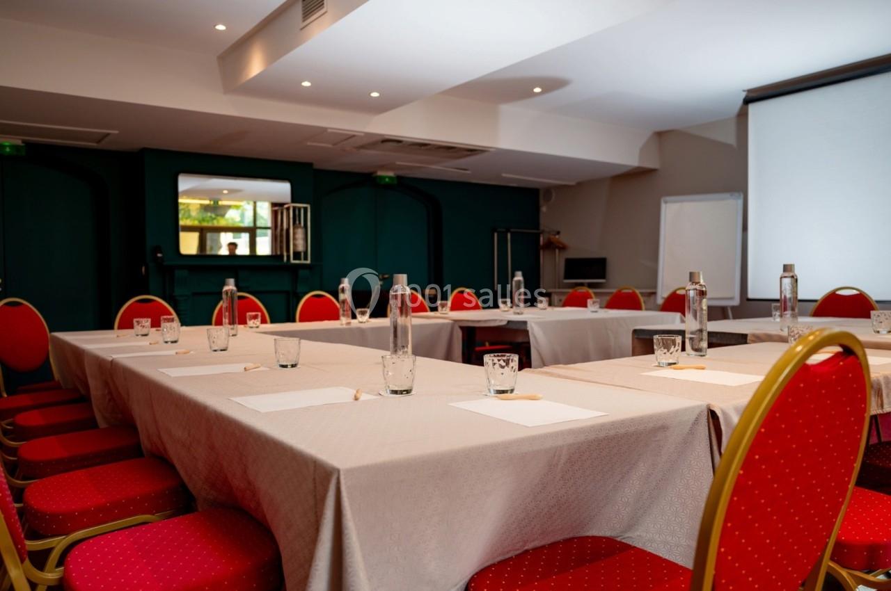 Salle de réunion avec tables en U, chaises rouges, bouteilles d'eau, verres, bloc-notes et écran de projection.