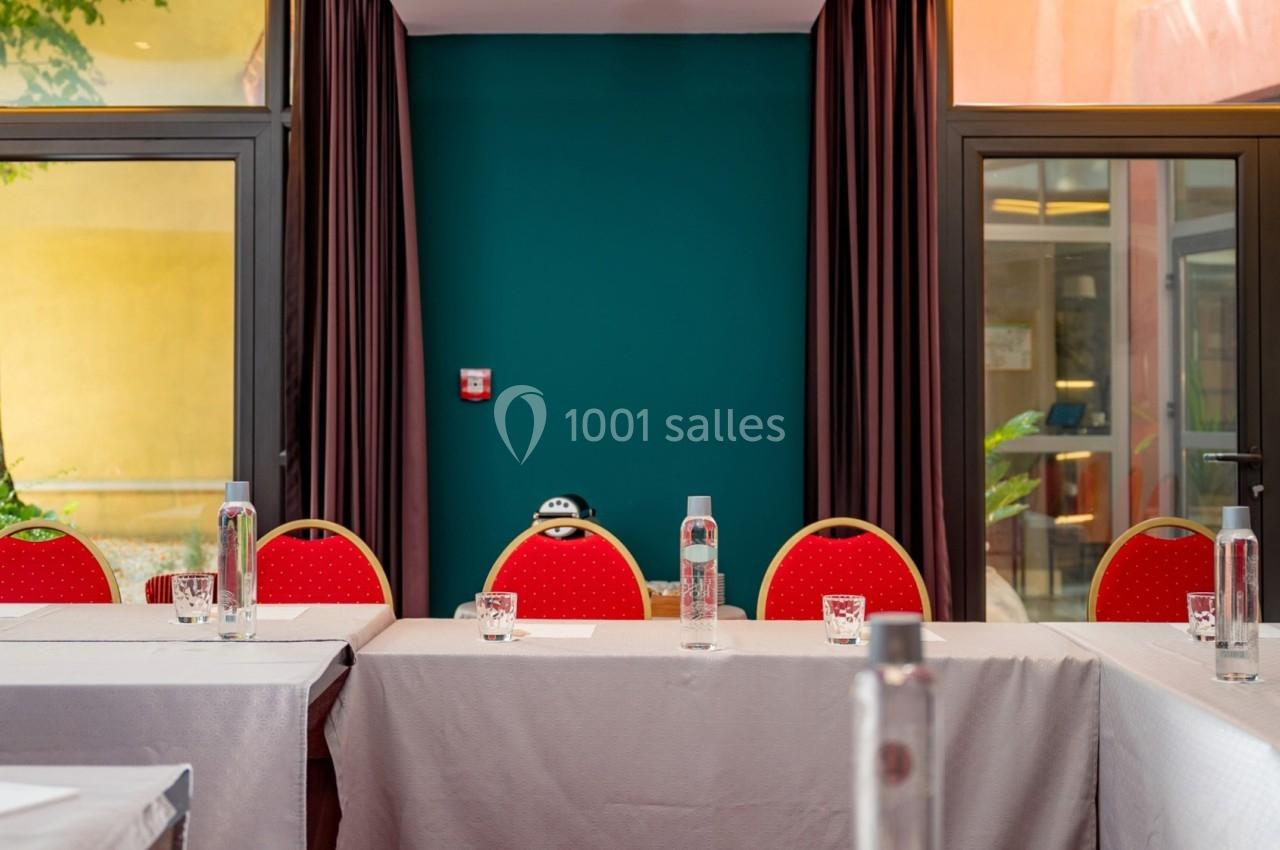 Salle de réunion avec tables recouvertes de nappes blanches, chaises rouges et bouteilles d'eau alignées.