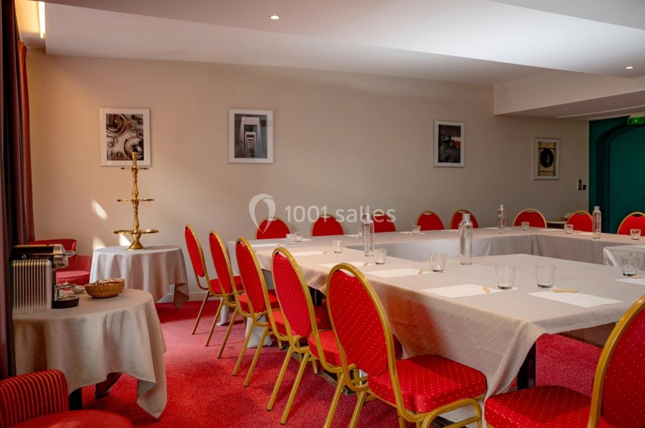 Salle de réunion avec tables disposées en U, chaises rouges, nappes blanches et décoration murale sobre.