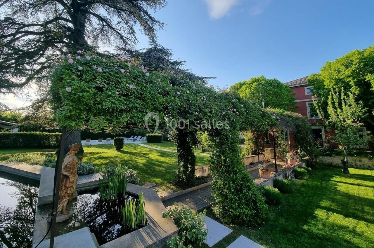 Pergola couverte de plantes grimpantes dans un jardin verdoyant avec bassin, pelouse et bâtiment en arrière-plan.