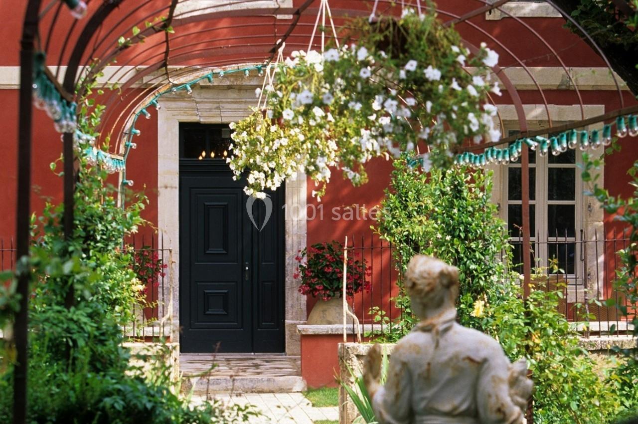 Entrée d'une maison rouge avec porte noire, encadrée par des fleurs suspendues et un jardin verdoyant au premier plan.