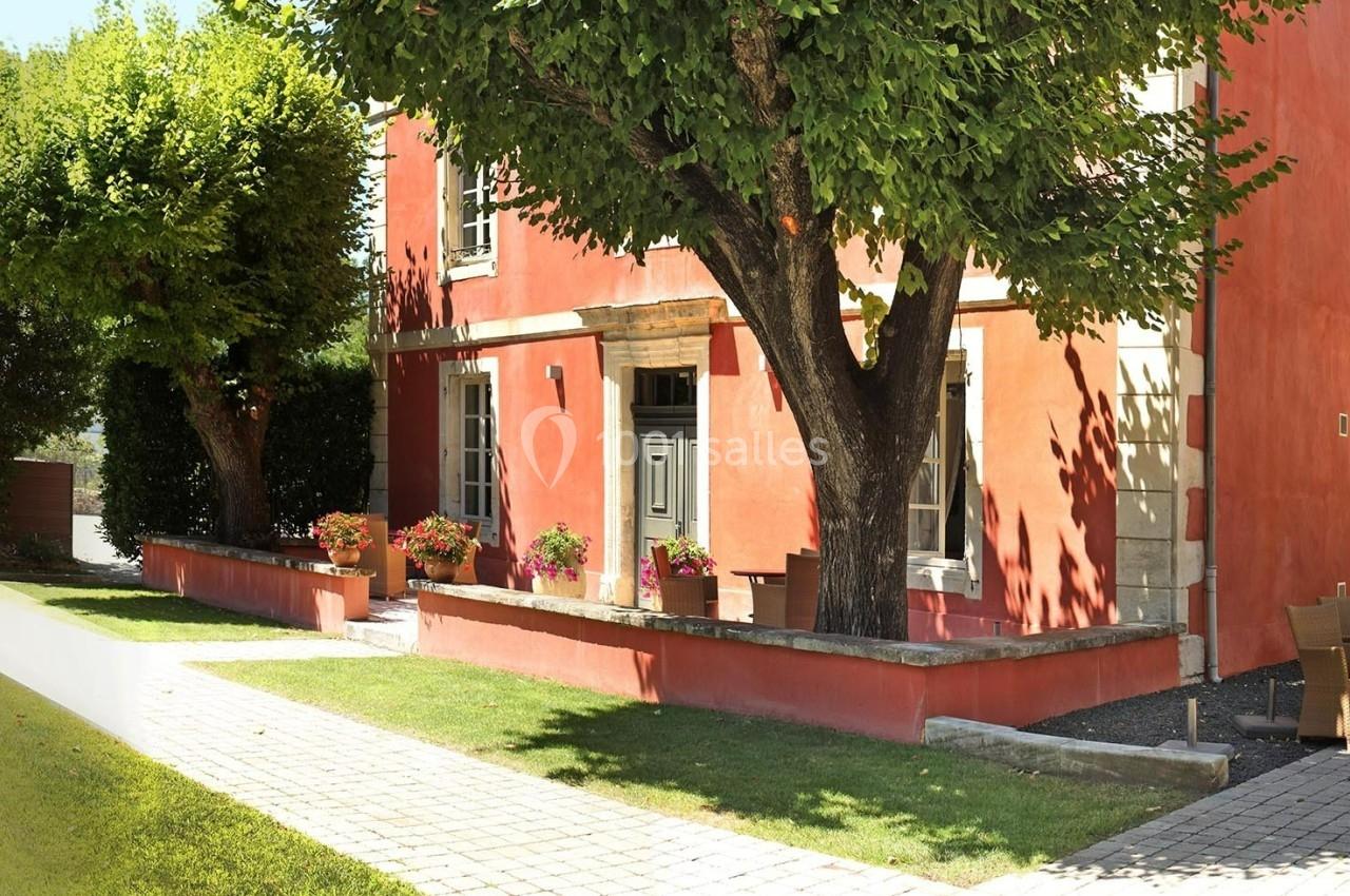 Façade d'une maison rouge avec des arbres, des jardinières fleuries et une allée pavée bordée de pelouse.