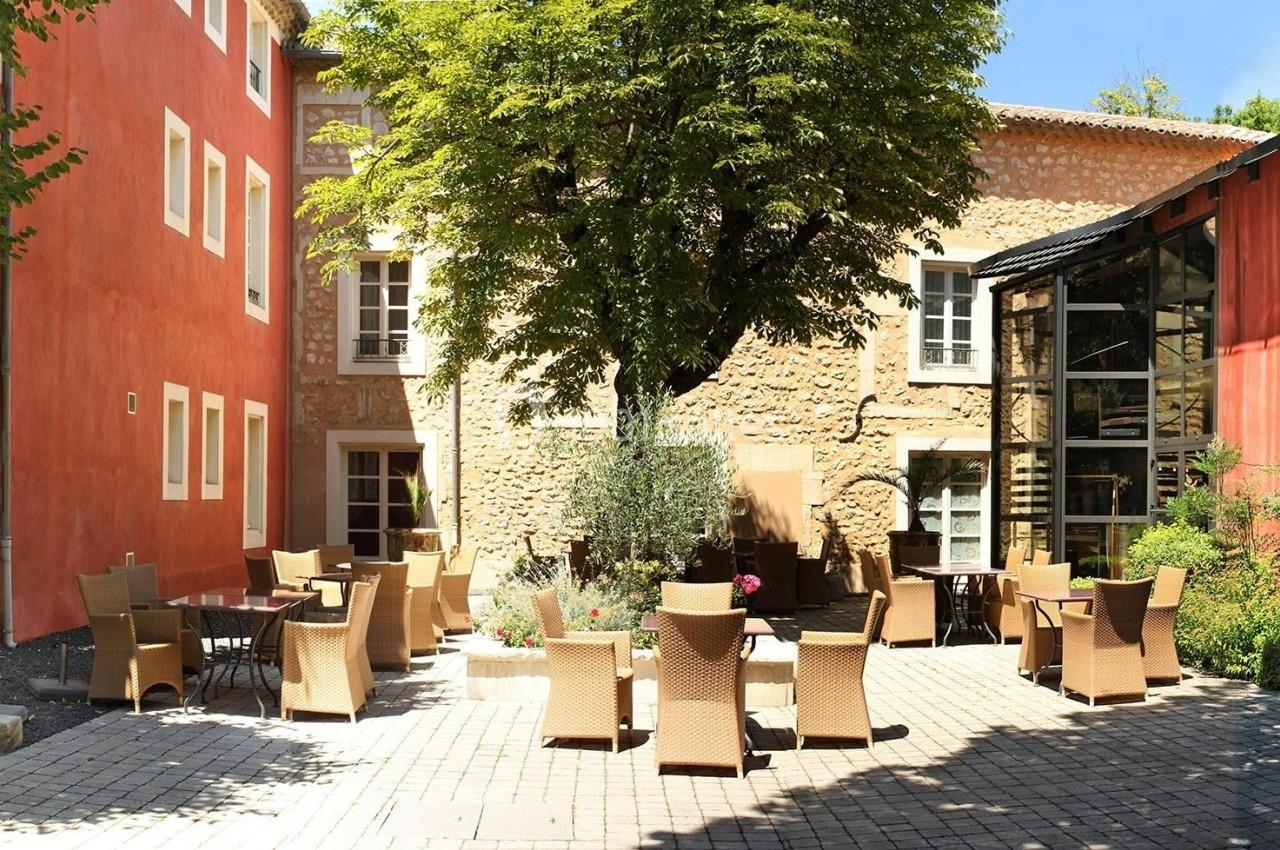Cour pavée avec des chaises et tables en rotin, entourée de bâtiments aux façades colorées et d'un arbre central.