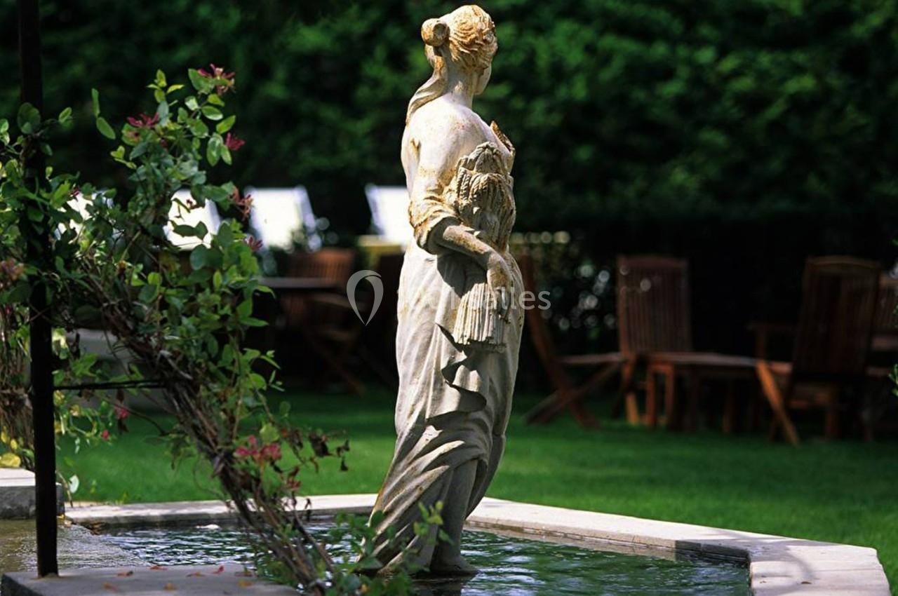 Statue en pierre représentant une femme tenant des épis de blé, placée au centre d'un bassin dans un jardin verdoyant.