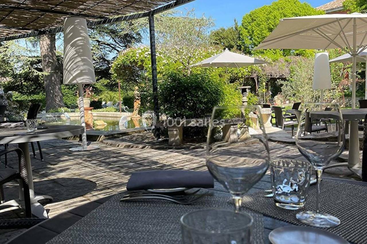 Terrasse ombragée avec tables dressées, vue sur un jardin verdoyant et parasols blancs par une journée ensoleillée.